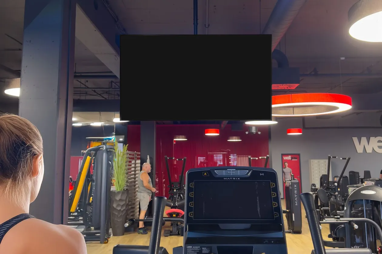 Ein Bildschirm über den Cardiogeräten eines Fitnessstudios, im Vordergrund eine Frau auf dem Crosstrainer