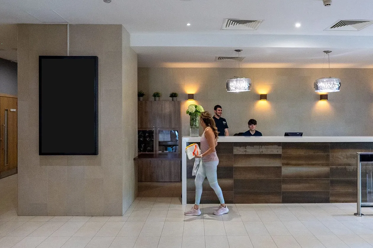 Ein Hochformat-Bildschirm an einer Säule neben dem Empfang eines Fitnessstudios, ein Mitglied geht vorbei