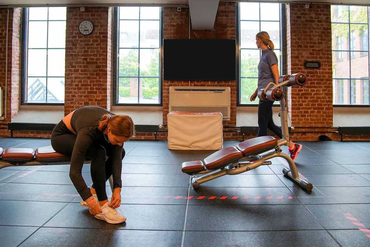 Ein Bildschirm an einer Backsteinwand zwischen hohen Fenstern in einem Fitnessstudio, eine Frau bindet sich auf einer Bank die Schuhe